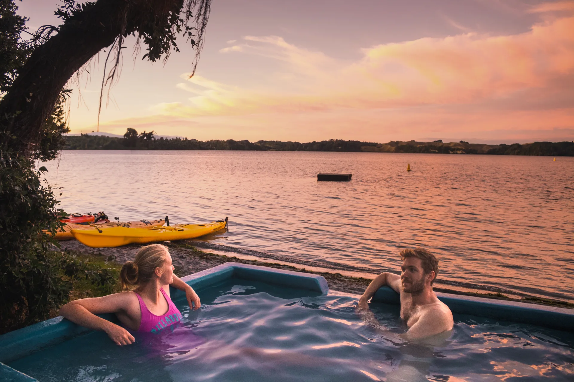 Two guests relax in a lakeside hot pool as the sun sets over the water