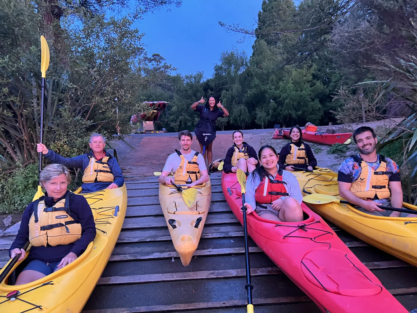 A group of guests in kayaks at dusk, with their guide behind them giving two thumbs up
