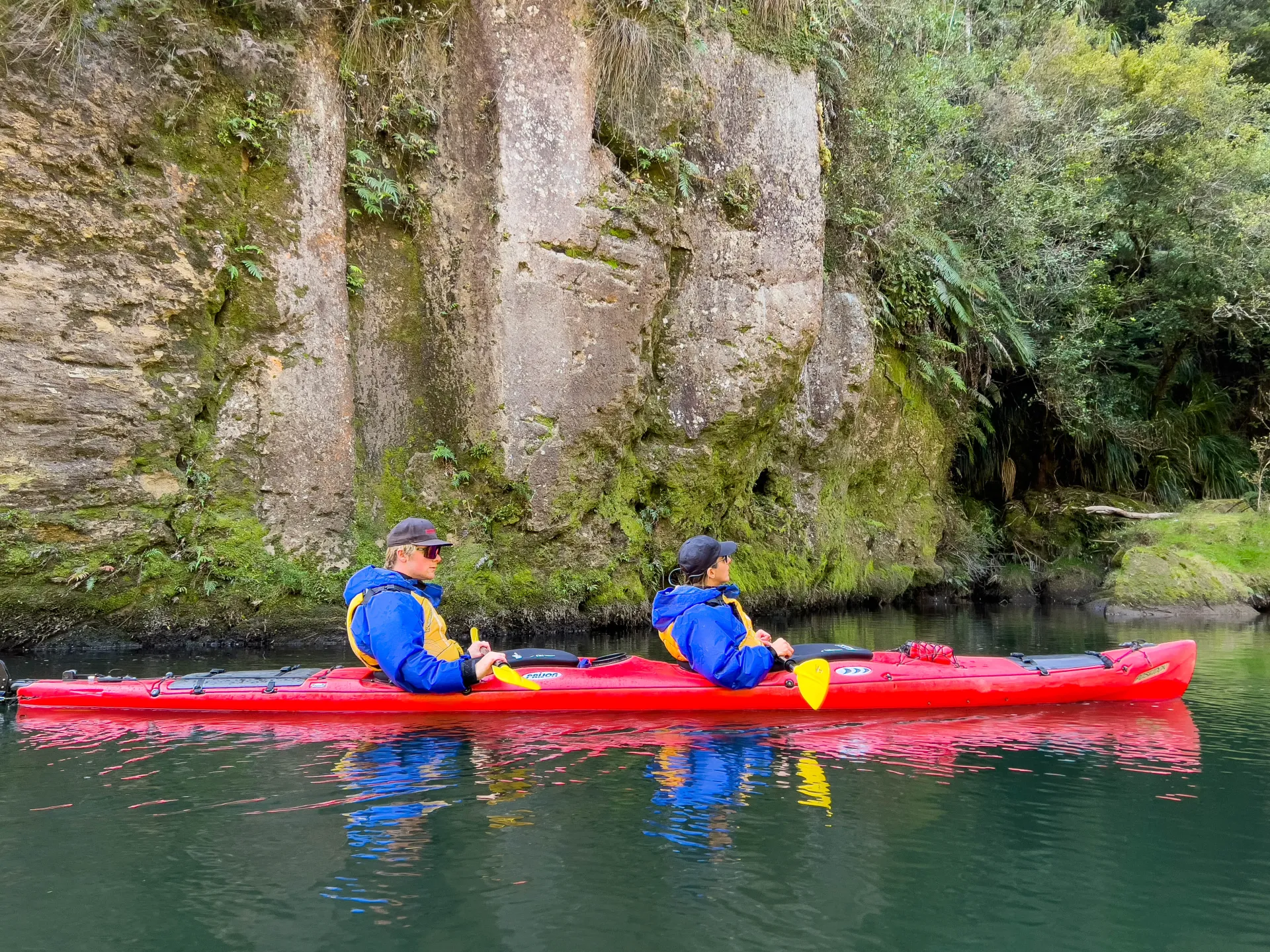 Two kayakers in a red kayak paddling past mossy canyon walls on Lake McLaren, Tauranga
